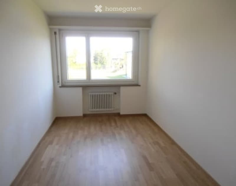 Empty room with wooden floor, white walls, large window, radiator