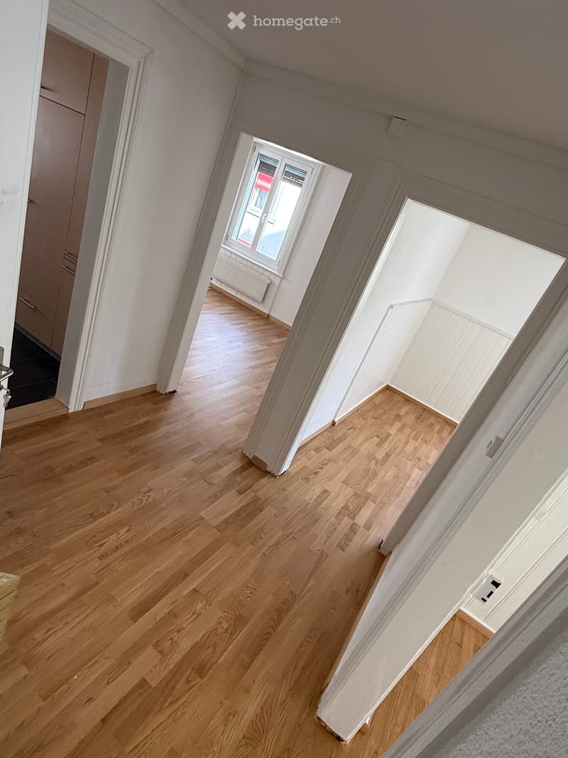 Empty room with wooden floors and white walls. There are three doorways leading to other rooms. One doorway has a window with a white frame and a white radiator below it.
