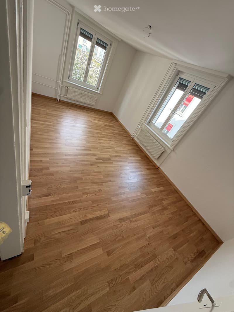 Empty room with wooden floors, white walls, and two windows with white frames. There are two white radiators under the windows.