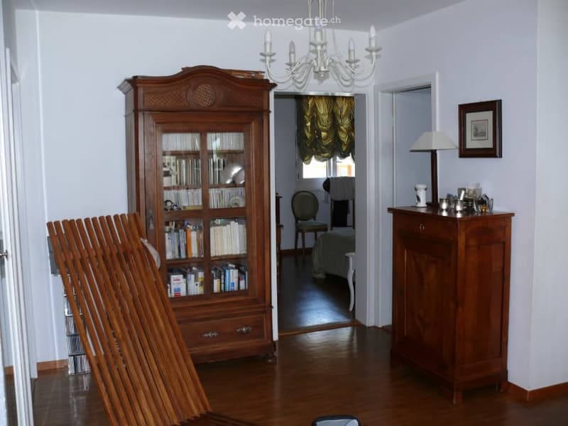 wooden floor, white walls, wooden cabinet with glass doors, wooden chair, chandelier, wooden cabinet with drawers, lamp, picture frame, books, stack of books
