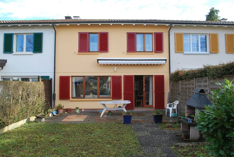 Three-story house with red shutters, white awning, and a garden with a table, chairs, and potted plants.