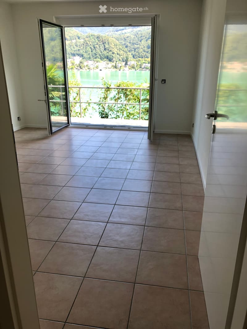 Empty room with tiled flooring, open sliding glass doors leading to a balcony with a scenic view of a lake and mountains.