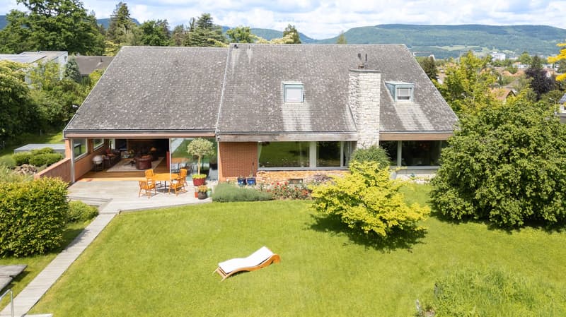 Detached house with gray roof, brick walls, glass windows, wooden deck, garden with lounger, outdoor table and chairs