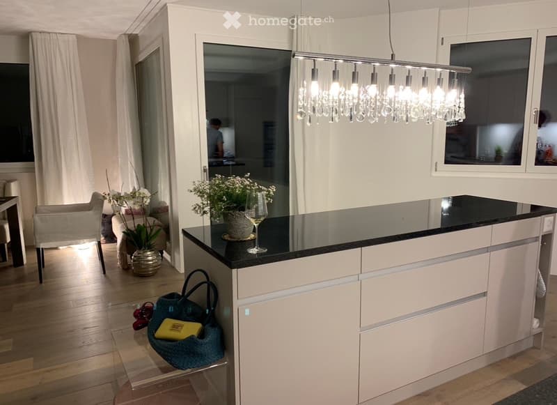 Kitchen. Modern kitchen with a black countertop, white cabinets, and a chandelier. A vase with flowers and wine glasses is on the counter. A chair and a table are in the background. A handbag and shoes are on the floor.