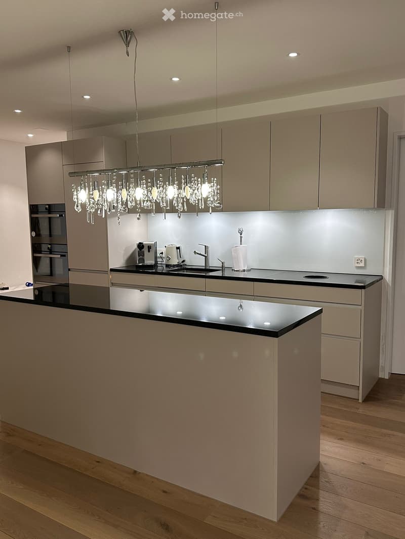 Kitchen. Modern kitchen with a large island, black countertops, and a chandelier. It includes a sink, coffee machine, toaster, and roll of paper towels. The cabinets are white, and the oven is built-in.