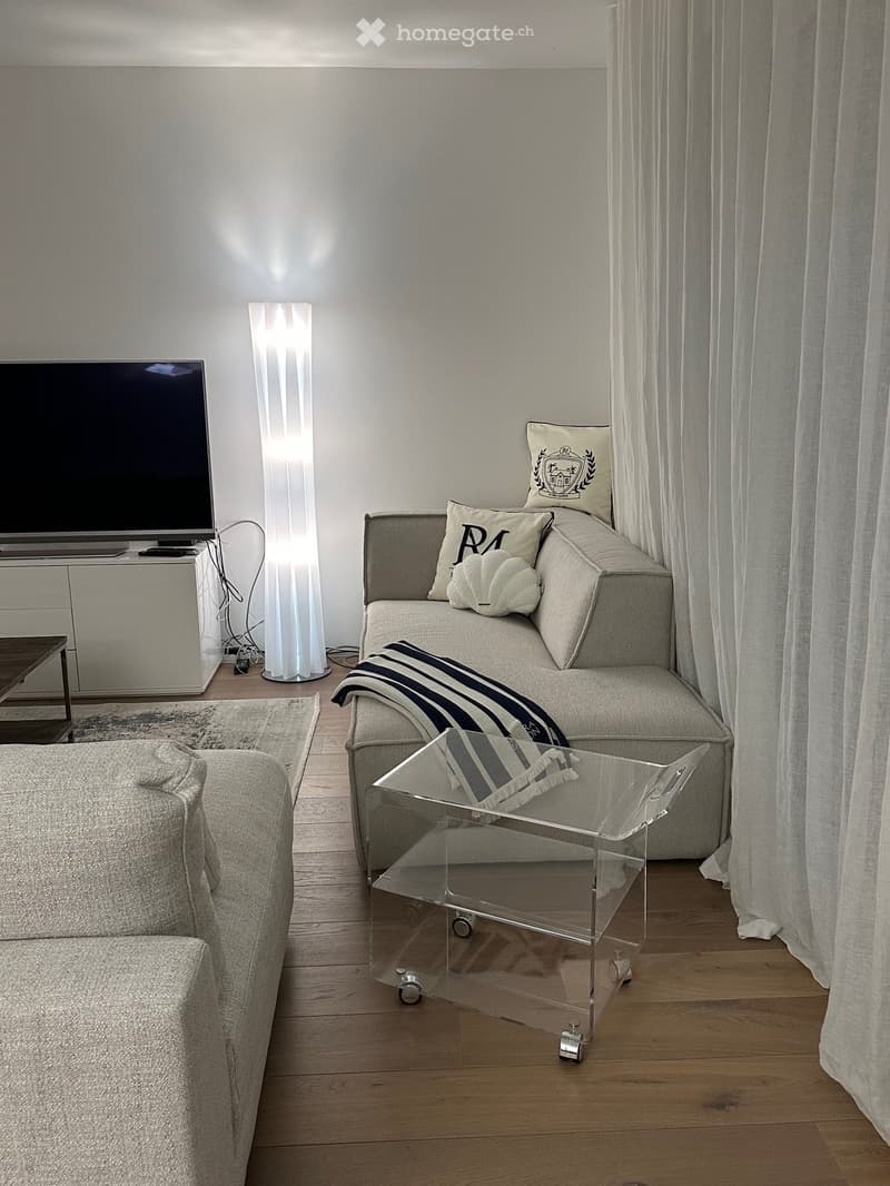 Furnished Room. Modern living room with a gray sofa, a clear glass coffee table, a floor lamp, and a TV on a white cabinet. The room has a wooden floor and white curtains.