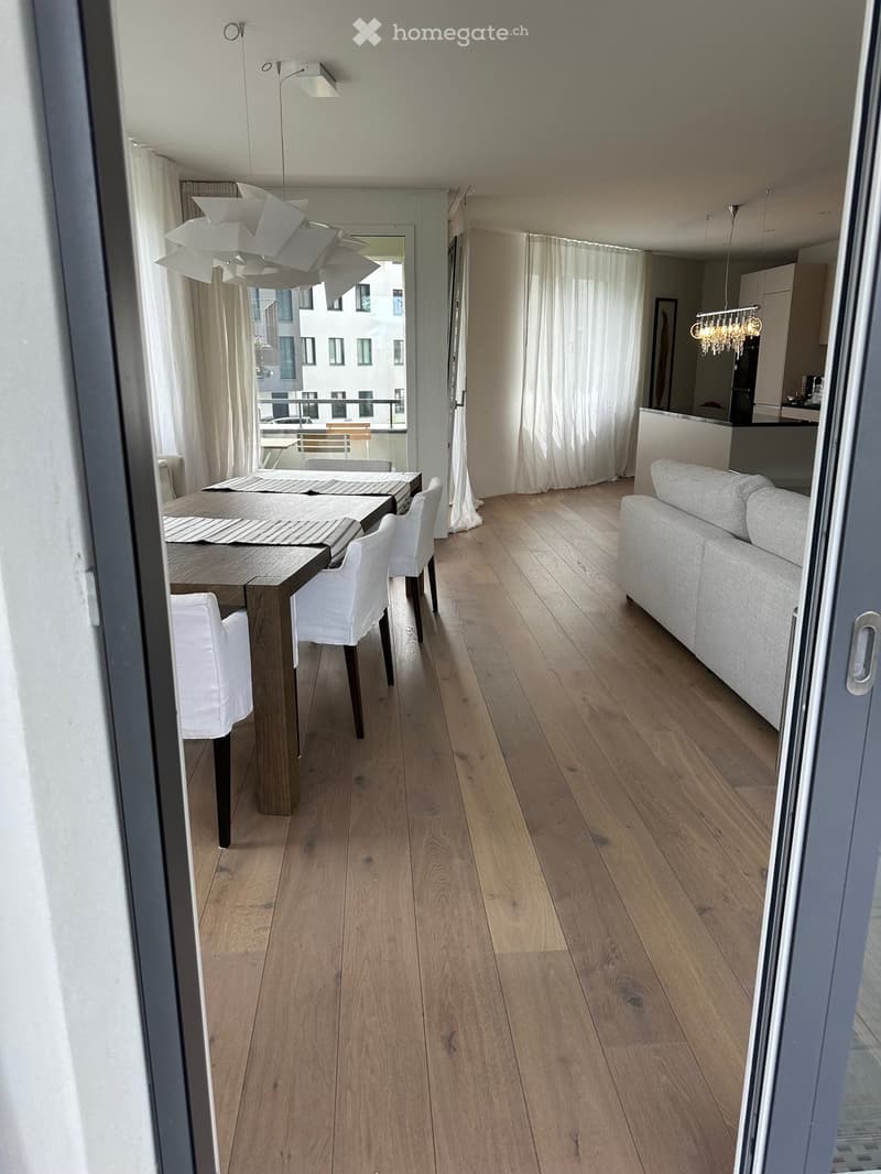 Furnished Room. Dining area with wooden table and chairs, a couch, and decorative lamps. The room has wooden floors and white curtains.