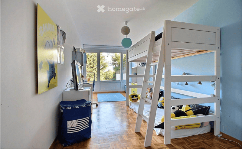 Bright bedroom with a bunk bed, desk, and a large window offering a view of greenery. The room features a blue rug, a colorful poster, and decorative pillows on the bunk bed.