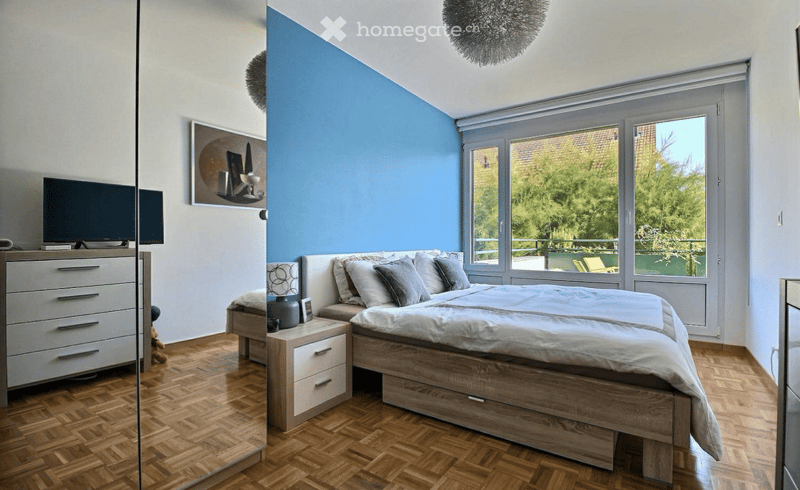 Spacious bedroom with wooden floor, blue accent wall, large bed, nightstand, dresser, and TV. Large windows with balcony access.