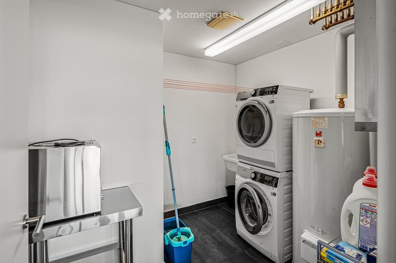 A small laundry room with a washing machine, dryer, and sink. A bucket and mop are placed on the floor. A water heater and detergent bottles are on the right side. A stainless steel table is on the left side.
