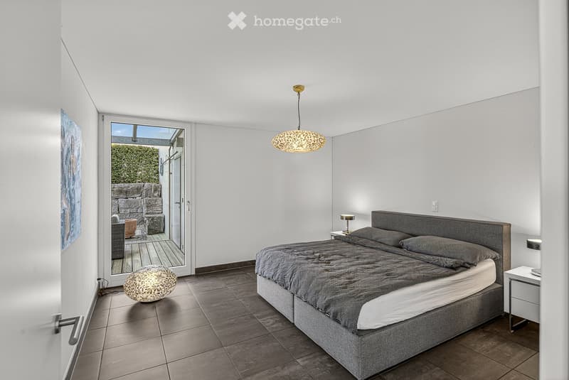 Modern bedroom with a double bed, gray bedspread, and pillows. A lamp is on each nightstand. A floor lamp is on the left side. A glass door leads to a patio with a wooden deck.