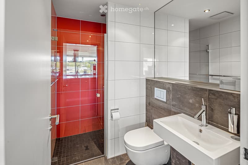 Modern bathroom with white tiles, red accent wall, glass shower door, white sink, and silver fixtures.