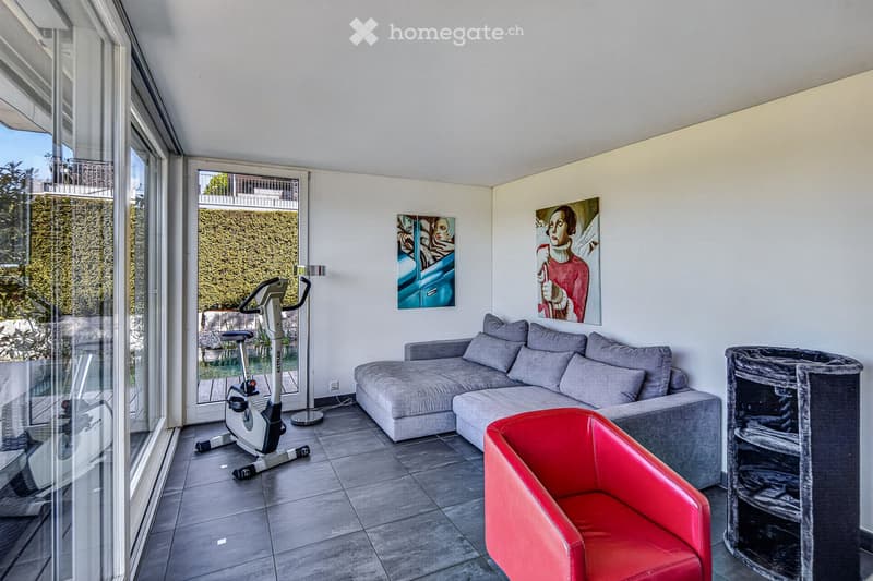 Modern furnished room with a couch, red chair, exercise bike, and cat tower. The room has large windows and a white ceiling.