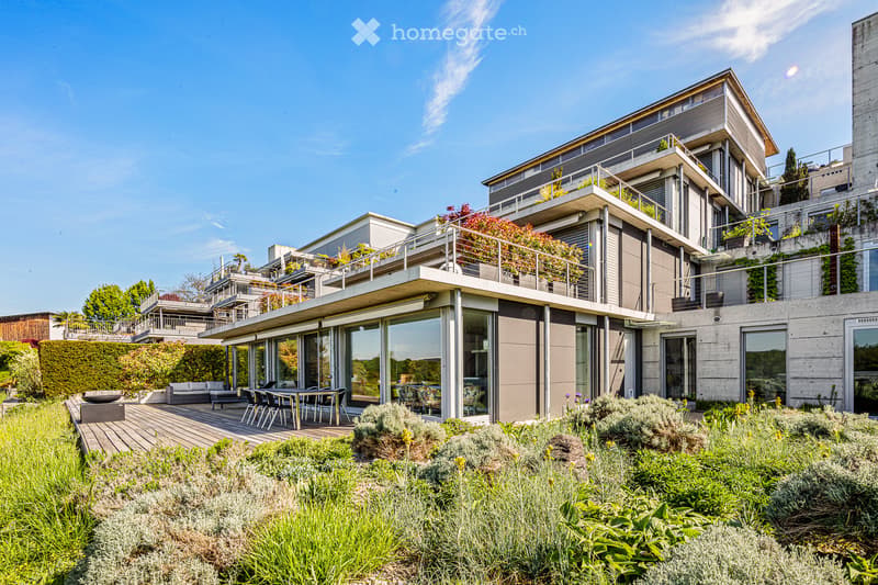 Modern multi-story building with balconies, large windows, and a raised ground floor. The exterior features a mix of concrete and glass, with green plants and a wooden deck.