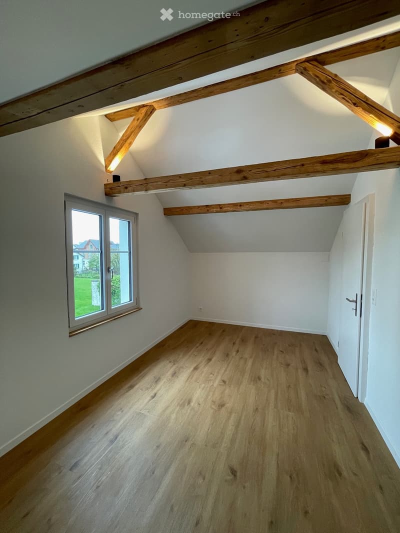 Empty room with wooden beams, white walls, wooden floor, and a window with a view of a grassy field.