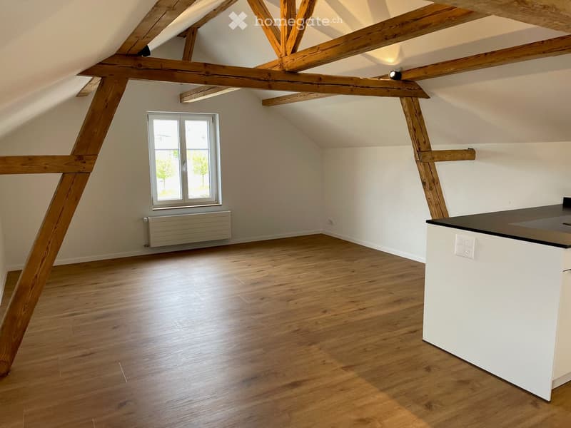 Empty attic room with wooden beams, wooden floor, and a window. A white radiator is mounted on the wall.