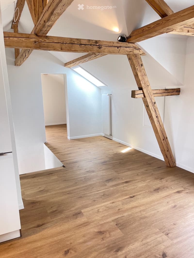 An empty attic room with wooden beams, wooden flooring, and white walls. There is a white radiator on the wall and a small window on the ceiling.