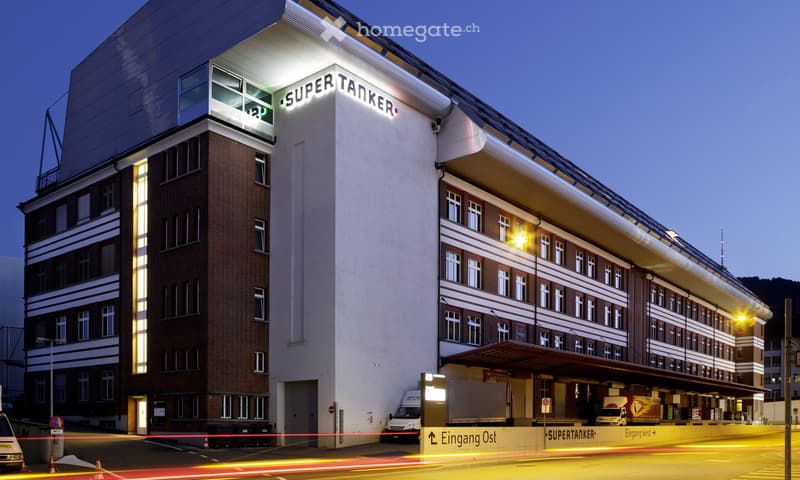 Modern office building with brick facade, glass windows, and a large sign reading 'SUPERTANKER'. The building has multiple floors, a flat roof, and a large entrance with a canopy.