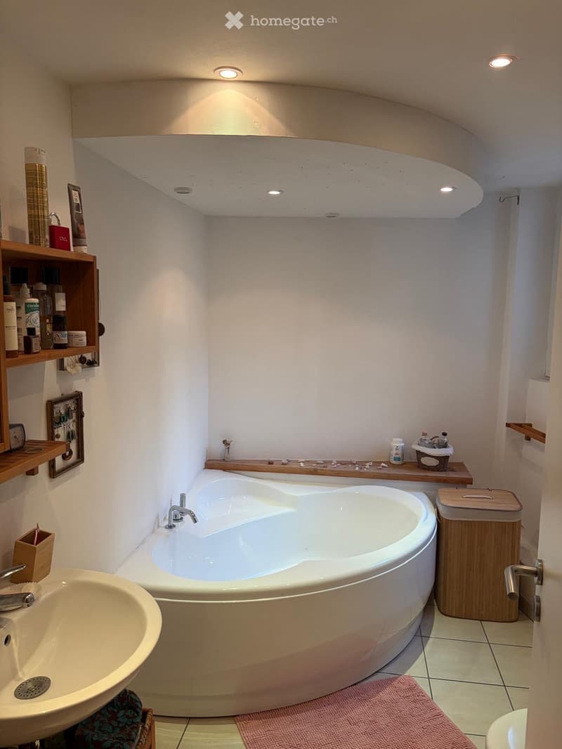 large bathtub, white sink, wooden shelves, tiled floor, pink rug, trash bin, wooden door