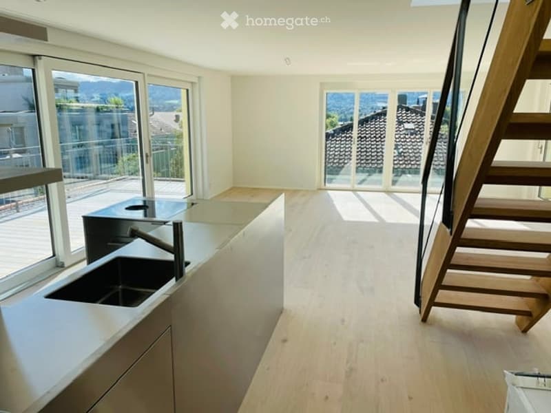Modern kitchen with stainless steel countertops, a sink, and a faucet. The kitchen is empty and has large windows and a wooden staircase.