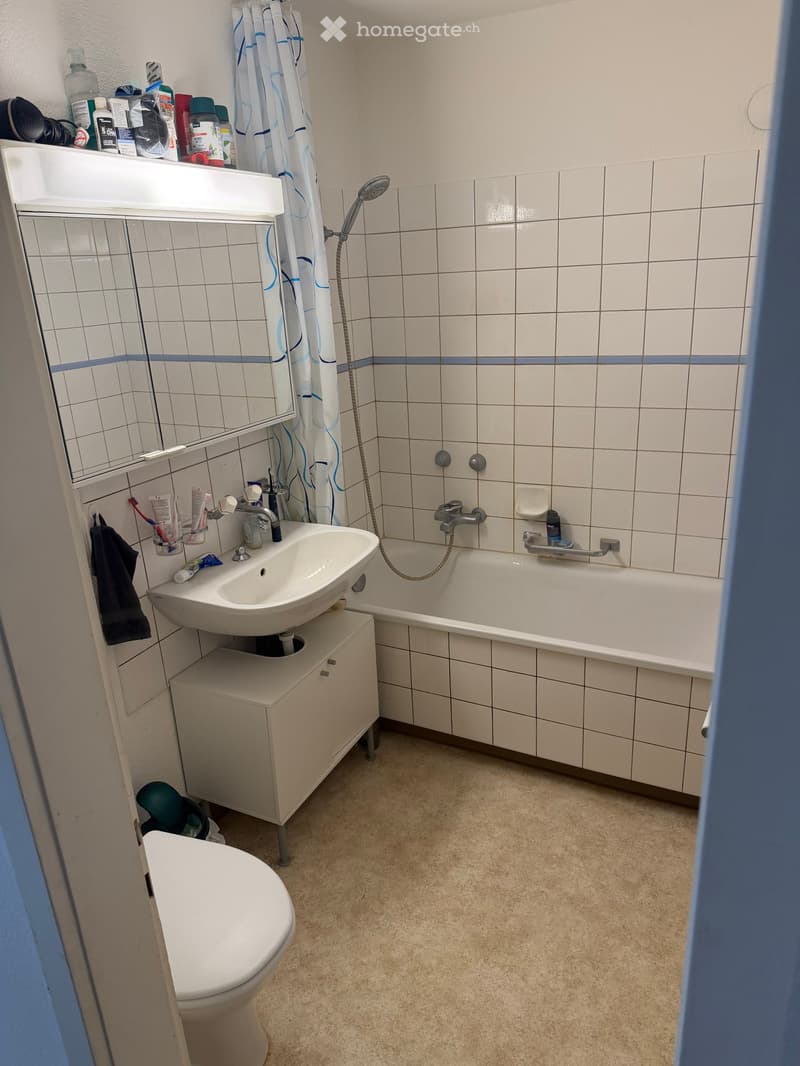 Bathroom with white tiled walls, a sink with a mirror, a bathtub with a shower curtain, a toilet, and a small cabinet under the sink.