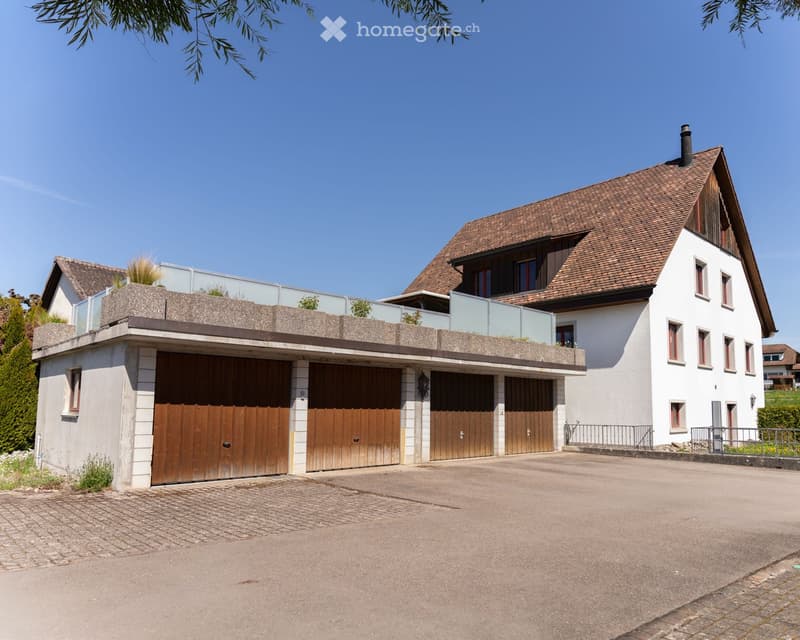 Two-story house with a brown tiled roof, white walls, multiple windows, and a large garage with wooden doors. The house features a modern balcony with a glass railing on the upper level.