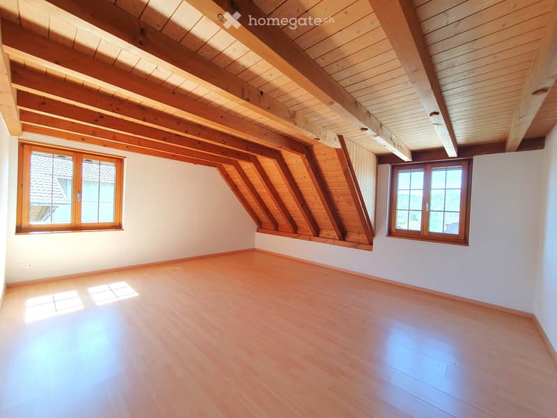 Empty attic room with wooden floors and exposed wooden beams on the ceiling. Two windows with wooden frames provide natural light.