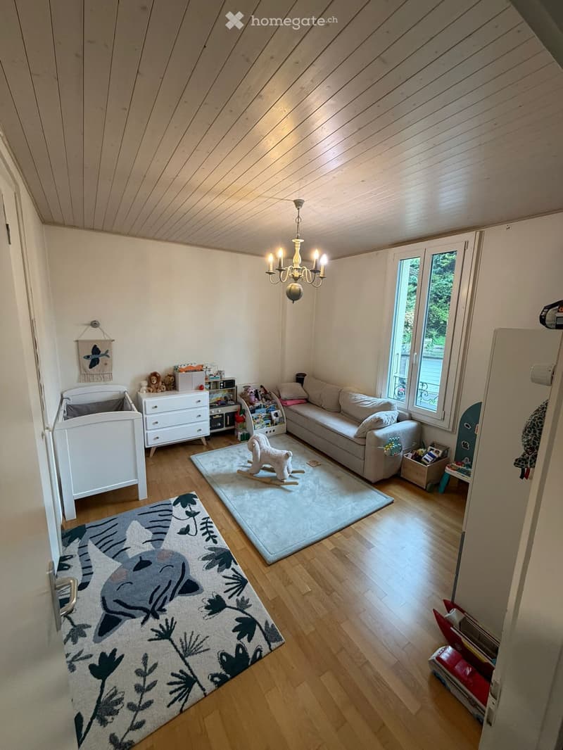 Bright, wooden-floored room with a slanted ceiling, featuring a sofa, rocking horse, baby crib, dresser, bookshelf, and chandelier.