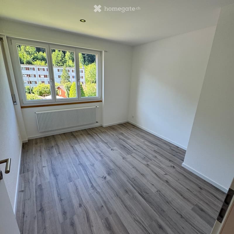 Empty room with wooden flooring, white walls, large window with view of trees and buildings, radiator, and a door handle visible.