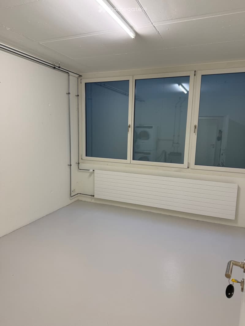 Empty room with white walls, white floor, large windows, white radiator, pipes on the wall, and a door.