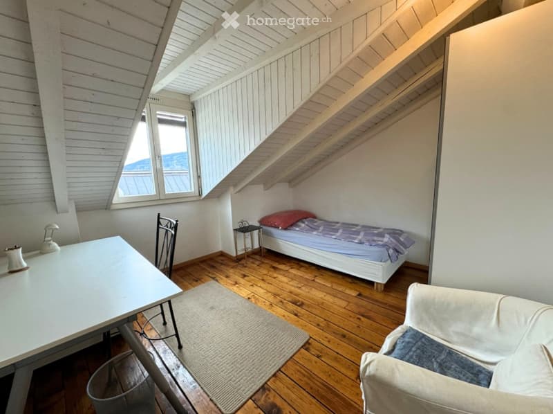 Small attic room with wooden floor, white walls, and wooden beams. Features a bed, desk, chair, and couch.