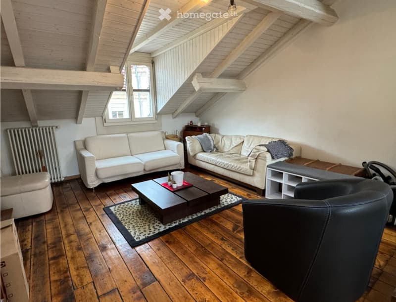 A furnished room with wooden floors, white walls, and a wooden ceiling. There are two white couches, a black leather chair, a coffee table, and a rug. A radiator is on the left wall, and a basket is on the window sill. A wooden box and shelves are on the