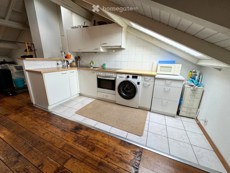 A small kitchen with white cabinets, wooden floor, and white tiles. It includes a washing machine, oven, sink, and a microwave. There is a rug on the floor and cleaning products on the counter.