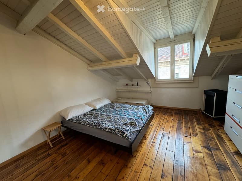 A bedroom with wooden floor, white walls, and wooden ceiling. A bed with pillows is placed in the center. A side table with a lamp is on the left. A cabinet and shelves are on the right. Cups and bottles are on the shelf. A window is on the wall.