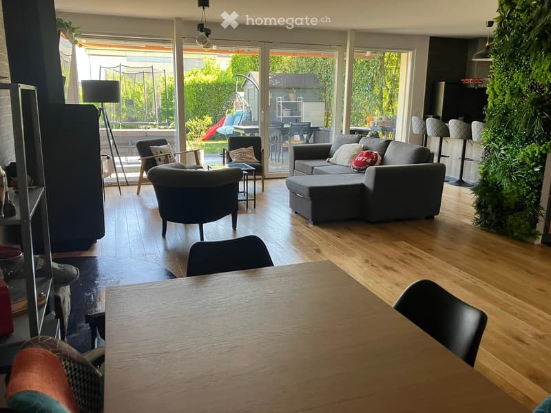 Furnished Room. Spacious living room with wooden floor, large windows, and sliding glass doors leading to a garden. A grey L-shaped sofa with pillows, a side table, and a lamp. A dining table with chairs. A plant wall and a swing in the garden.