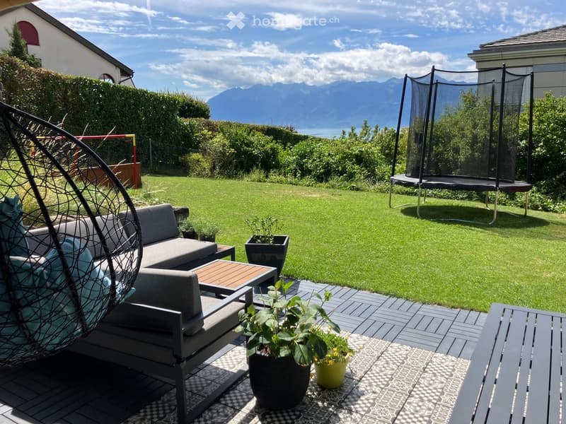Outdoor seating area with a couch, table, and a hammock. Potted plants and a trampoline are in the garden. The garden has a view of the mountains.