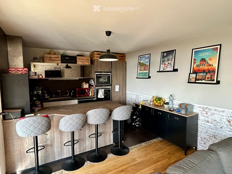 Kitchen. Modern kitchen with wooden island, bar stools, and a wine rack. Appliances include a refrigerator, microwave, and oven. The wall features brick accents and picture frames. A cabinet with a fruit bowl and bottle is on the counter. A chair is place