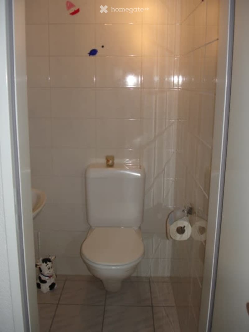 A small bathroom featuring a white toilet, a sink, a small toy cow, white tiles, and roll of toilet paper.