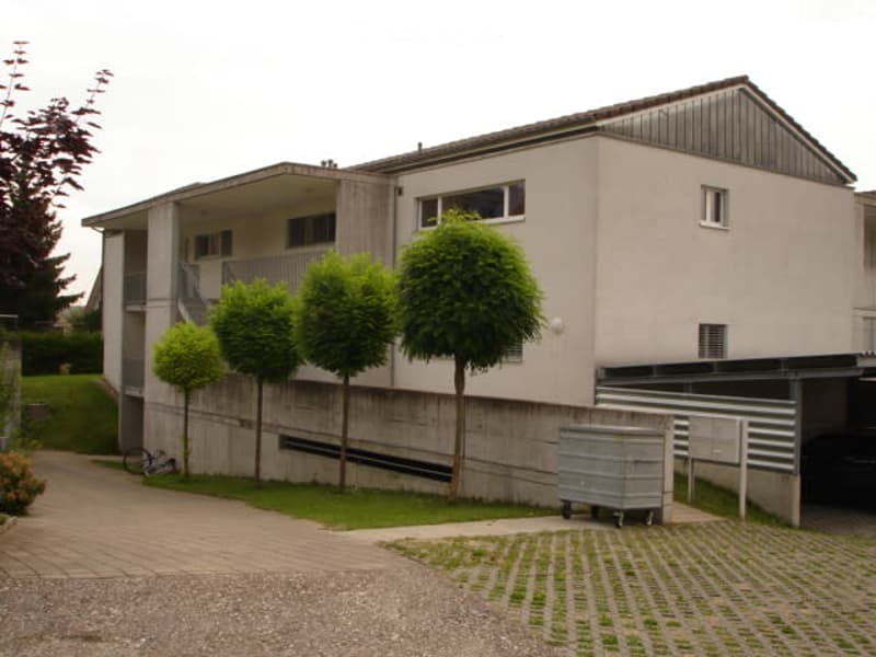 A modern two-story house with a sloped roof, white exterior, and a fenced entrance. It has several windows, a car parked in a garage under the building, and a balcony accessible via a staircase.