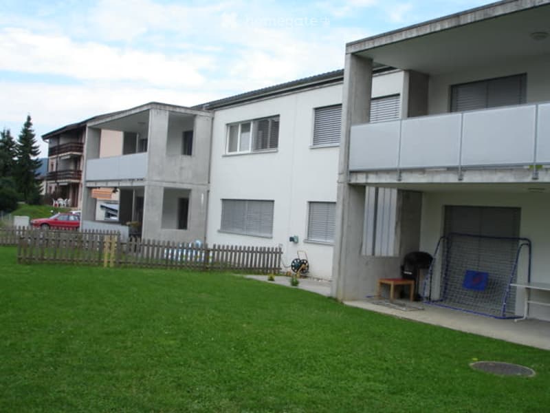 Condominium with balconies, white walls, metal fencing, red car parked, and green lawn.