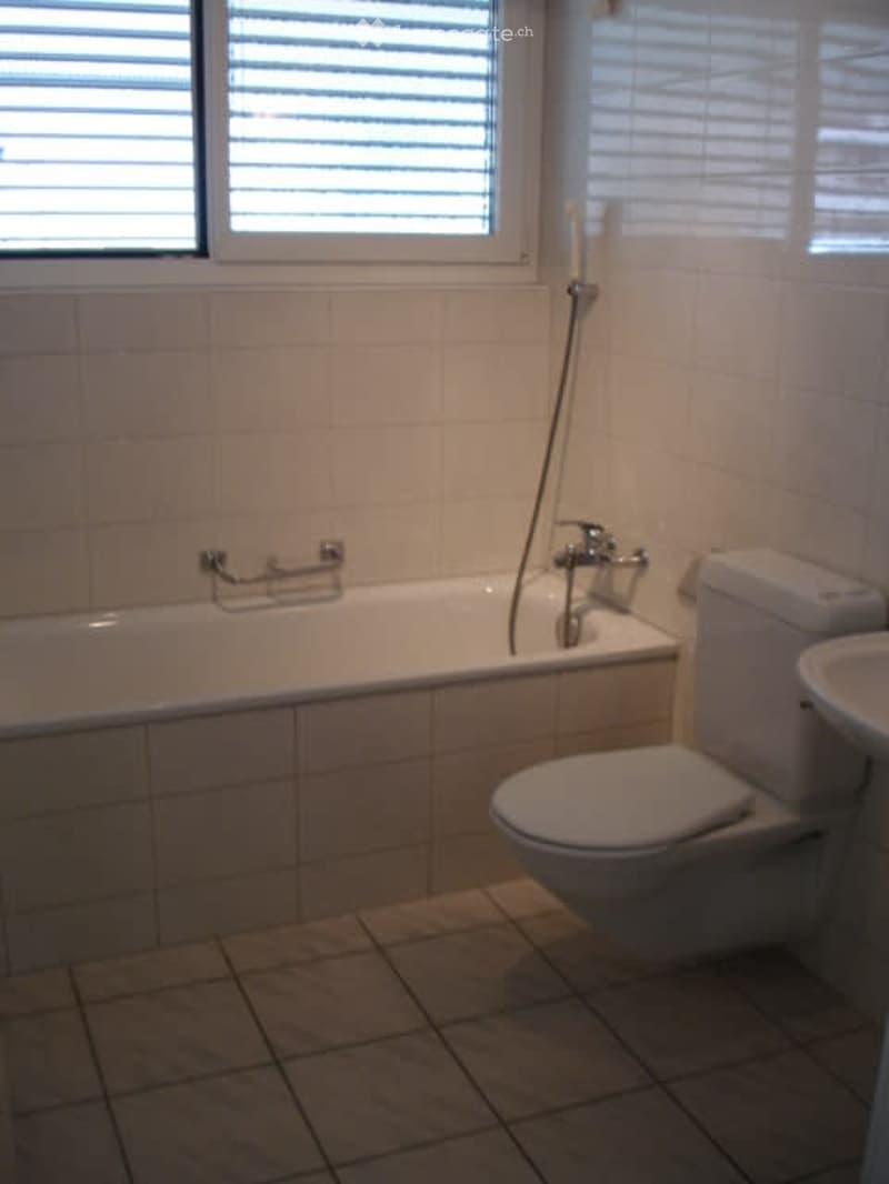 Bathroom with bathtub, toilet, sink, white tiles, silver fixtures, shower head, and a window with blinds.