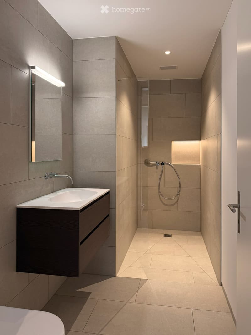 Modern bathroom with a white sink, mirror, and wooden cabinet. The shower area is separated by a glass wall, and the floor is tiled.