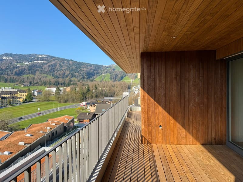 Wooden floor balcony with metal railing, wooden wall, and glass door, offering a view of the surrounding landscape.