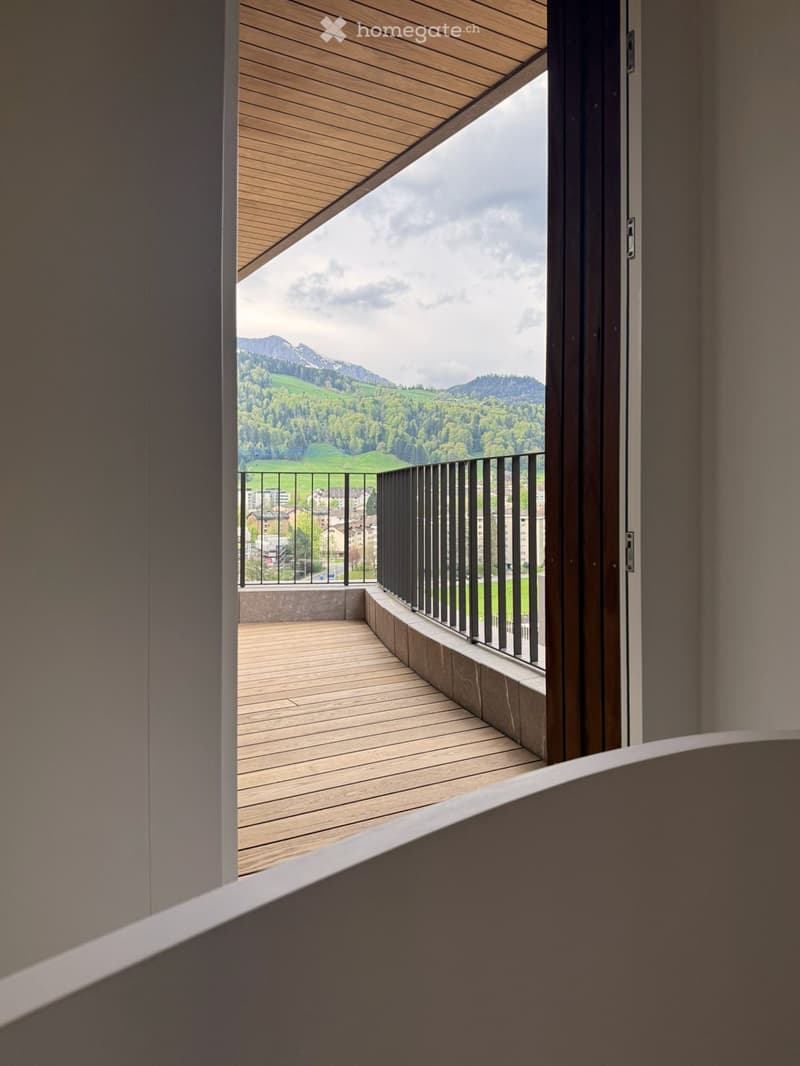 A balcony with a wooden floor and a black railing, offering a view of a valley with mountains and a town in the distance.