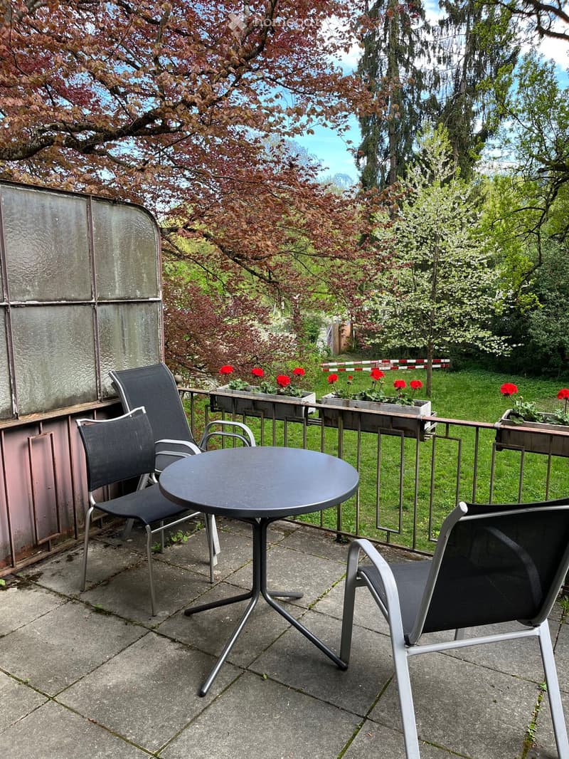 Outdoor balcony with a table and chairs, surrounded by a metal railing, and a garden with trees and flowers.