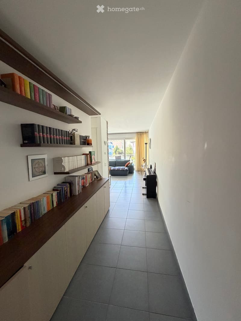 A hallway with tiled flooring, white walls, and built-in wooden shelves filled with books. A picture frame is mounted on the wall. A piano is placed in the hallway. A couch with pillows is in the living room. A plant and a lamp are on the side table. A ru