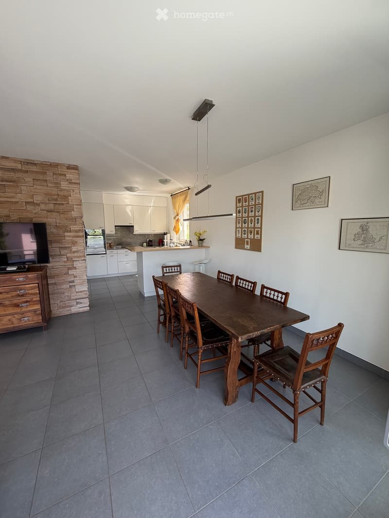 Dining area with a large wooden table and chairs, a TV on a cabinet, and a kitchen in the background.