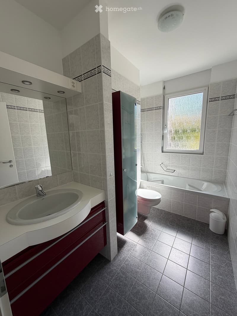 Bathroom with a large mirror, a sink, a toilet, a bathtub, and a shower door. The walls and floor are tiled. There is a trash can and a light fixture on the ceiling.