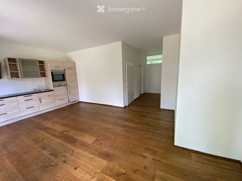 Empty room with wooden floors, white walls, kitchen area with cabinets, oven, and sink, and a window.