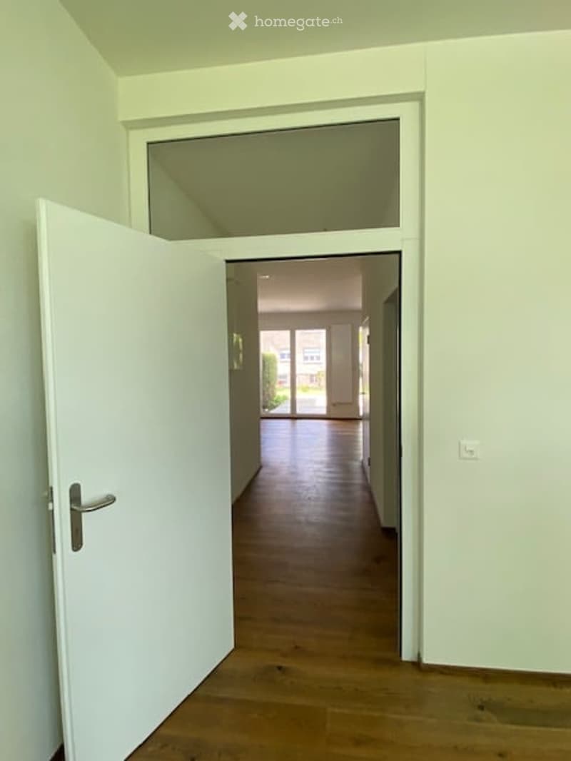 Empty hallway with wooden floor, white walls, open door, glass window, and a glimpse of a garden outside